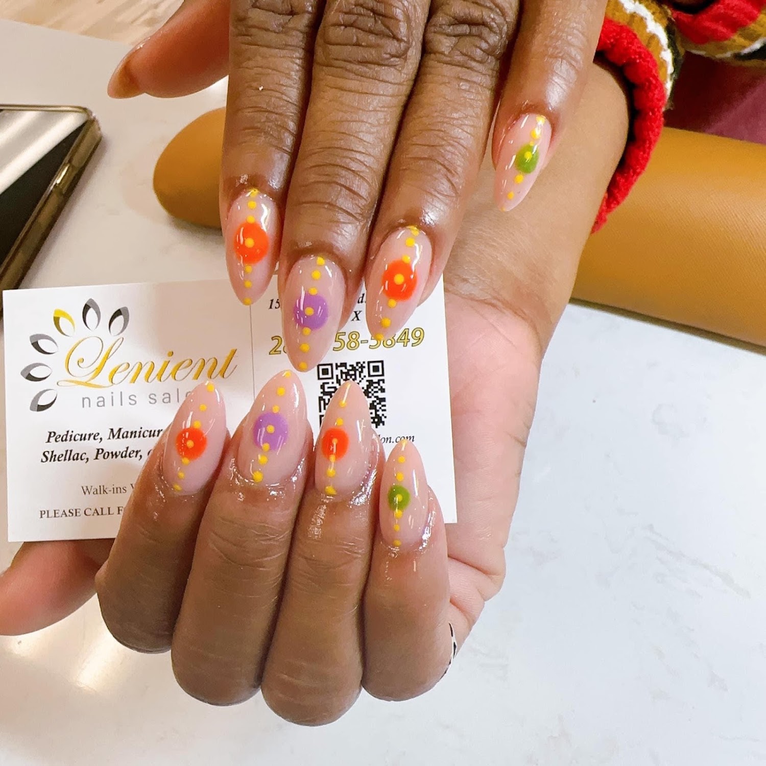 A client's hands at Lenient Nails holding the salon's business card, nails finished with orange, violet and green dots on nude polish.